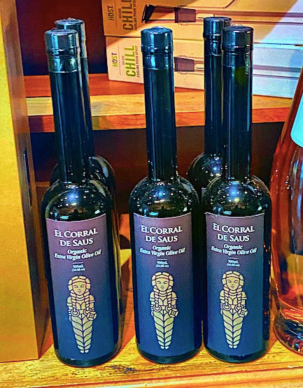 Three bottles of 'El Corral de Saus' olive oil on a wooden surface with a colorful background.